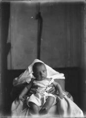 Portrait en intérieur d'un bébé tenu vers l'avant par deux mains féminines / [Anonyme].