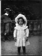 1 vue Portrait de petite fille en robe et chapeau blancs, un petit garçon à canotier à l'arrière plan. / [Anonyme]