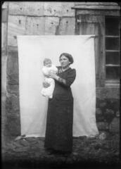 1 vue  - Portrait de femme tenant un enfant dans les bras devant une bâche blanche. / [SAUNAL, Léon (photographe)] (ouvre la visionneuse)