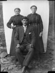 1 vue Portrait d'un homme assis sur une chaise et deux jeunes filles debout derrière lui, devant une bâche blanche. / [SAUNAL, Léon (photographe)]